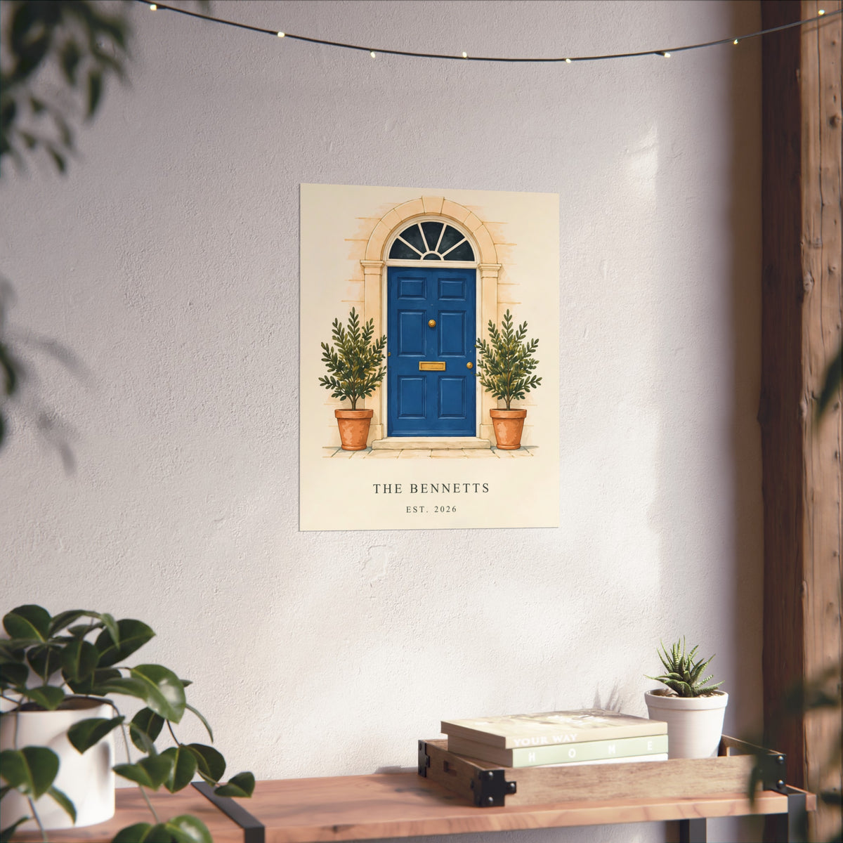 Our Little Door personalised art print with a blue front door, potted plants and example household wording, styled on a wall above a wooden console