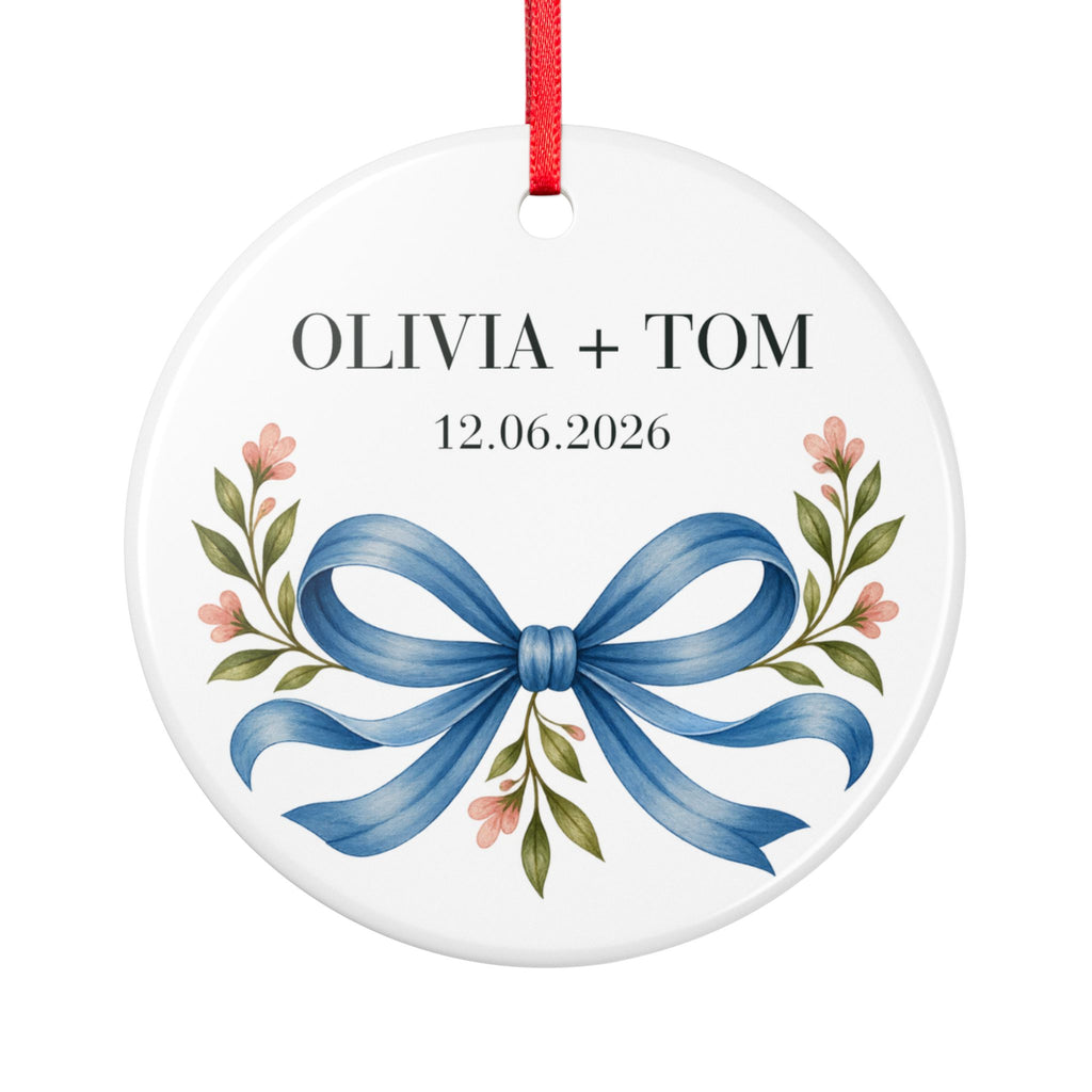 Round ceramic hanging ornament showing blue illustrated bow and pink flowers beneath sample names Olivia + Tom and a sample date.
