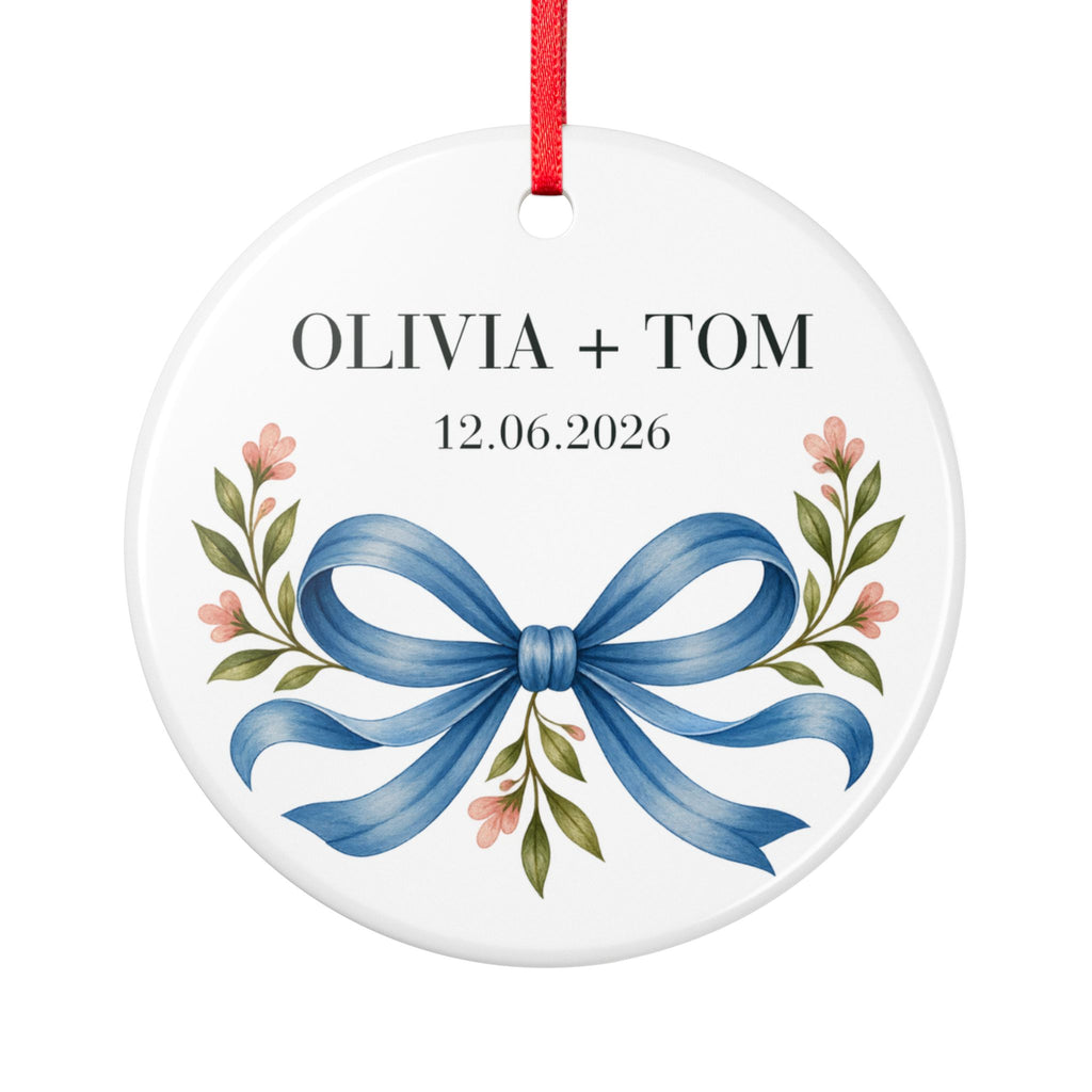 Round ceramic hanging ornament showing blue illustrated bow and pink flowers beneath sample names Olivia + Tom and a sample date.
