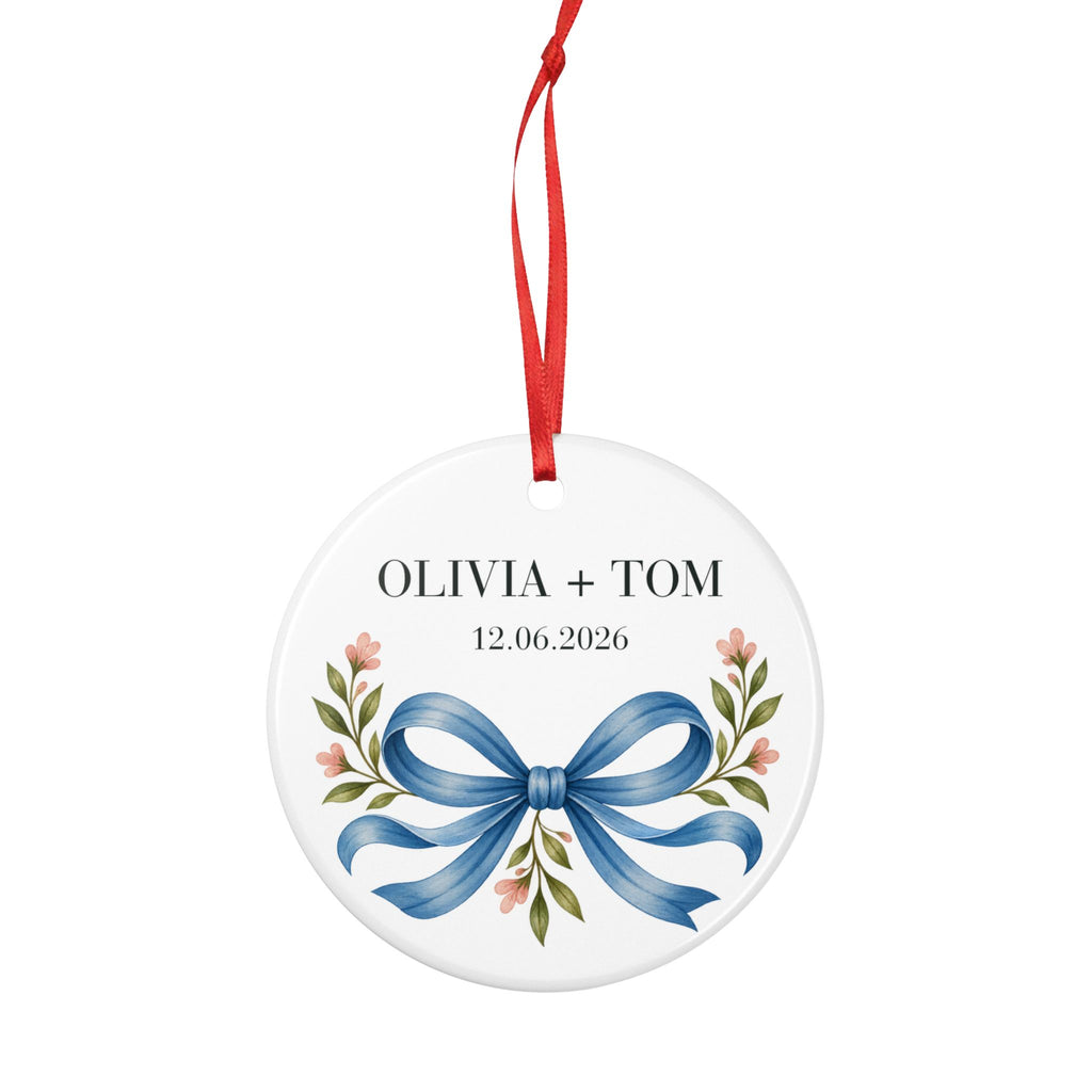 Round ceramic hanging ornament showing blue illustrated bow and pink flowers beneath sample names Olivia + Tom and a sample date.
