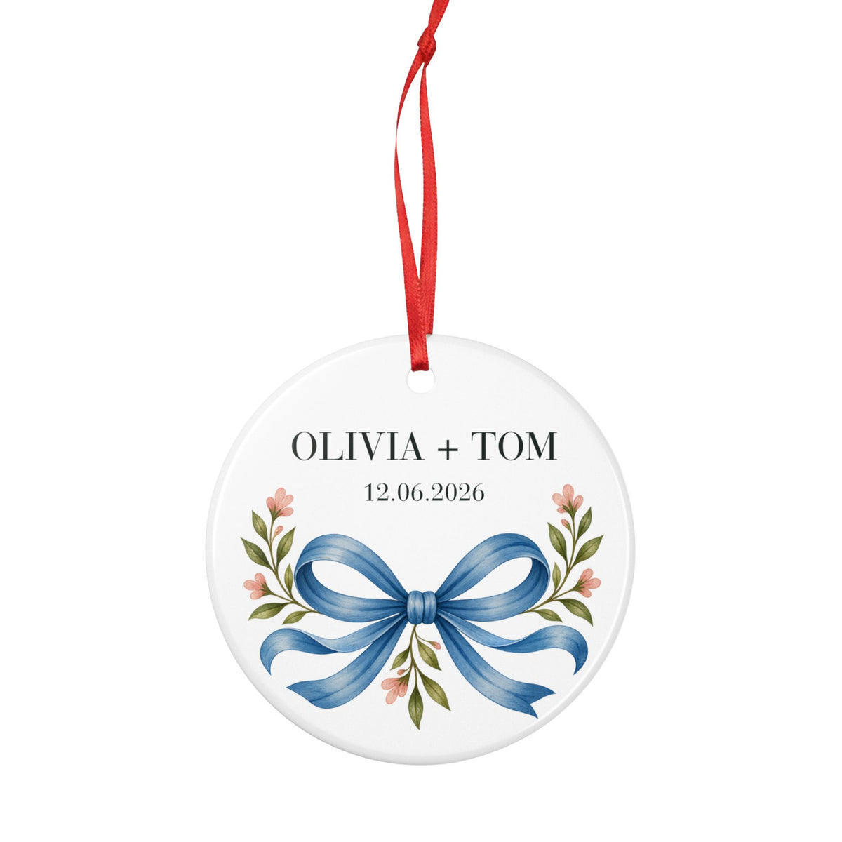 Round ceramic hanging ornament showing blue illustrated bow and pink flowers beneath sample names Olivia + Tom and a sample date.
