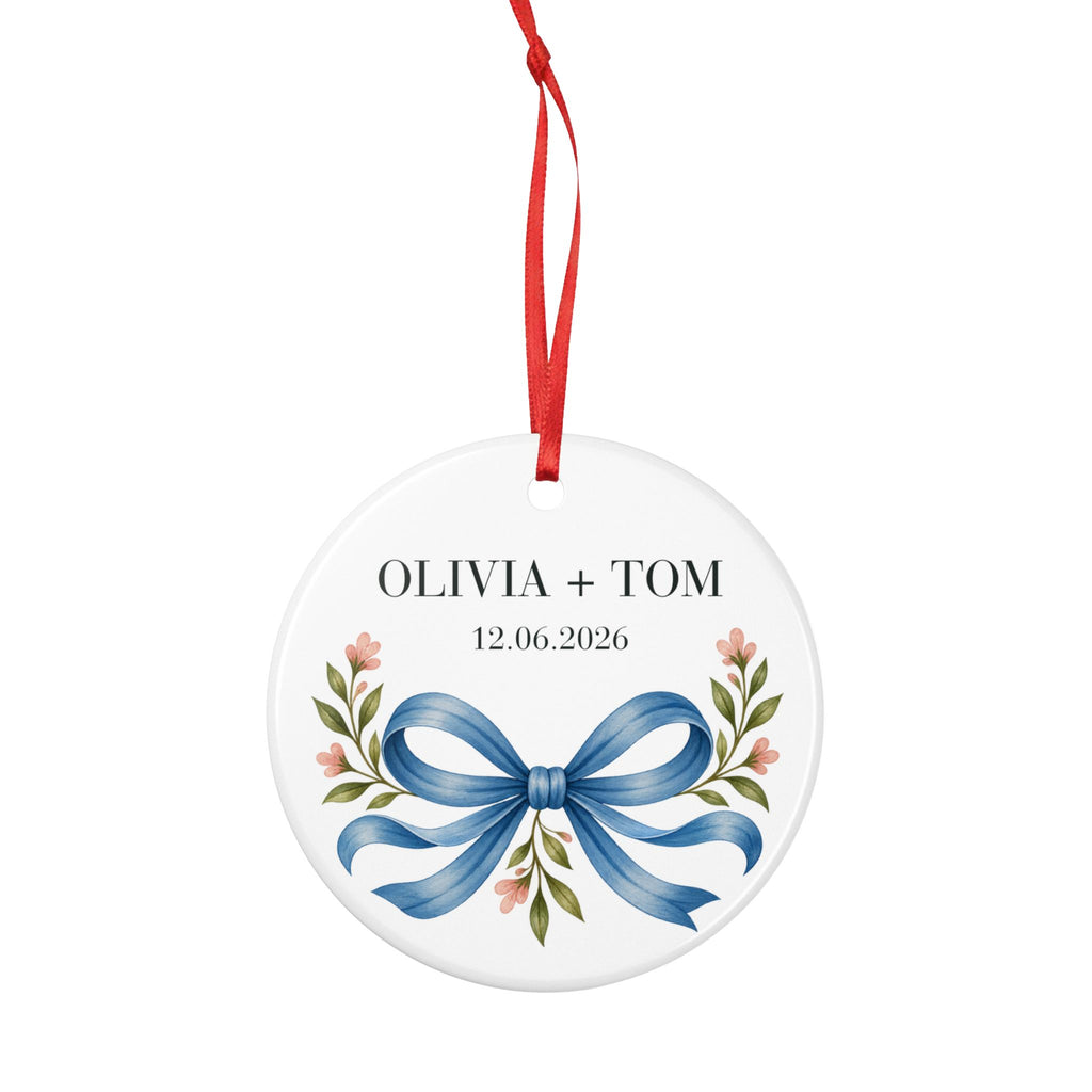 Round ceramic hanging ornament showing blue illustrated bow and pink flowers beneath sample names Olivia + Tom and a sample date.