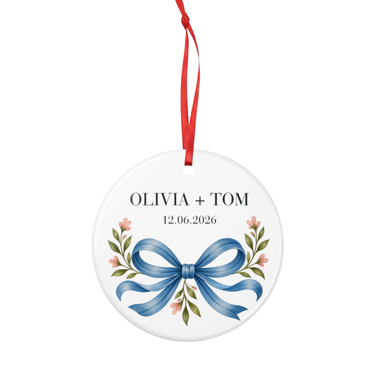 Round ceramic hanging ornament showing blue illustrated bow and pink flowers beneath sample names Olivia + Tom and a sample date.