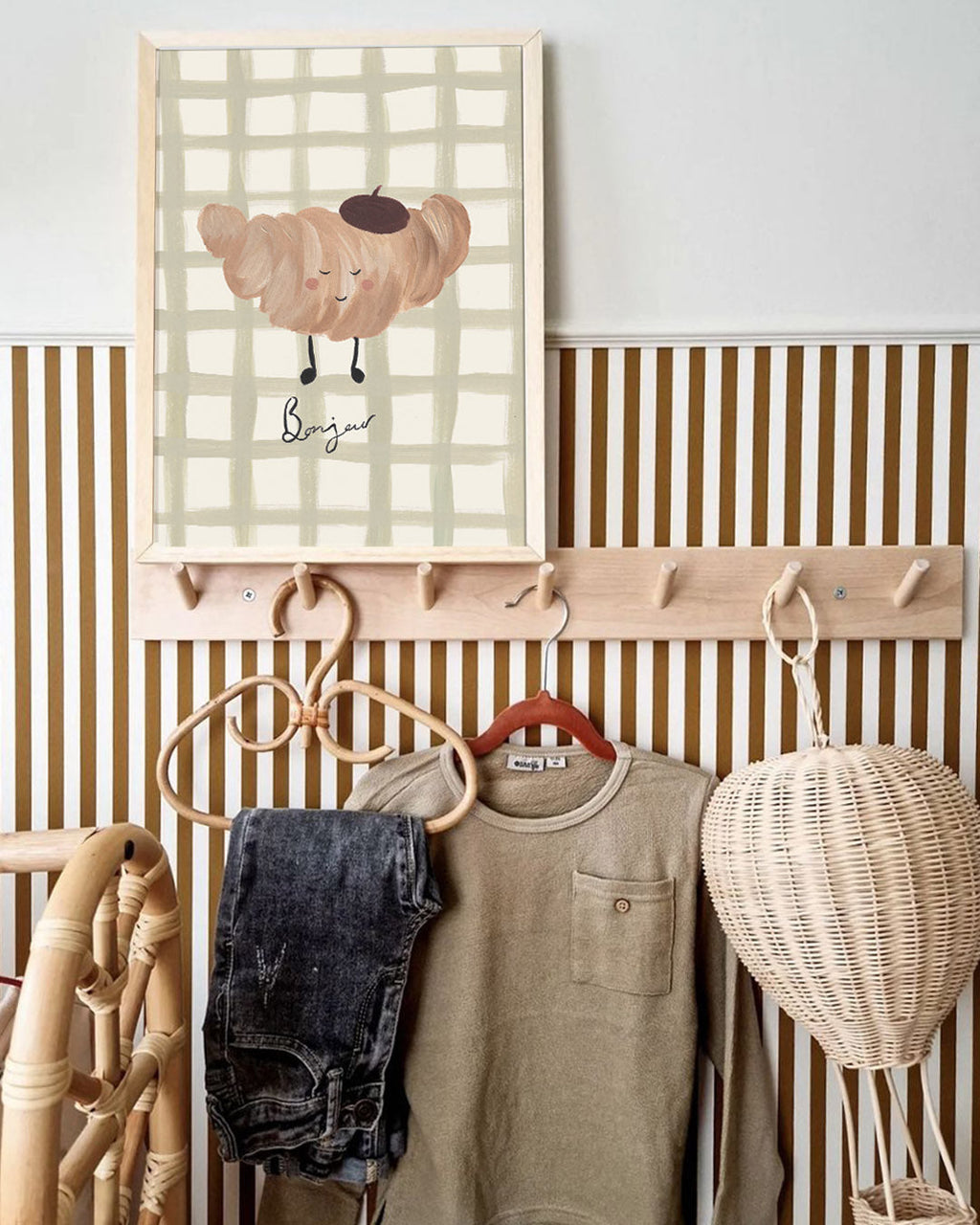 Croissant Bonjour print displayed above a nursery peg rail with children's clothes and a wicker bag.