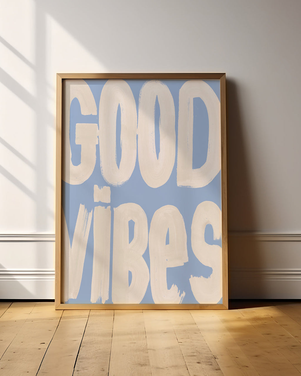 Good Vibes cream and blue typography print in a wooden frame on a sunlit wooden floor.