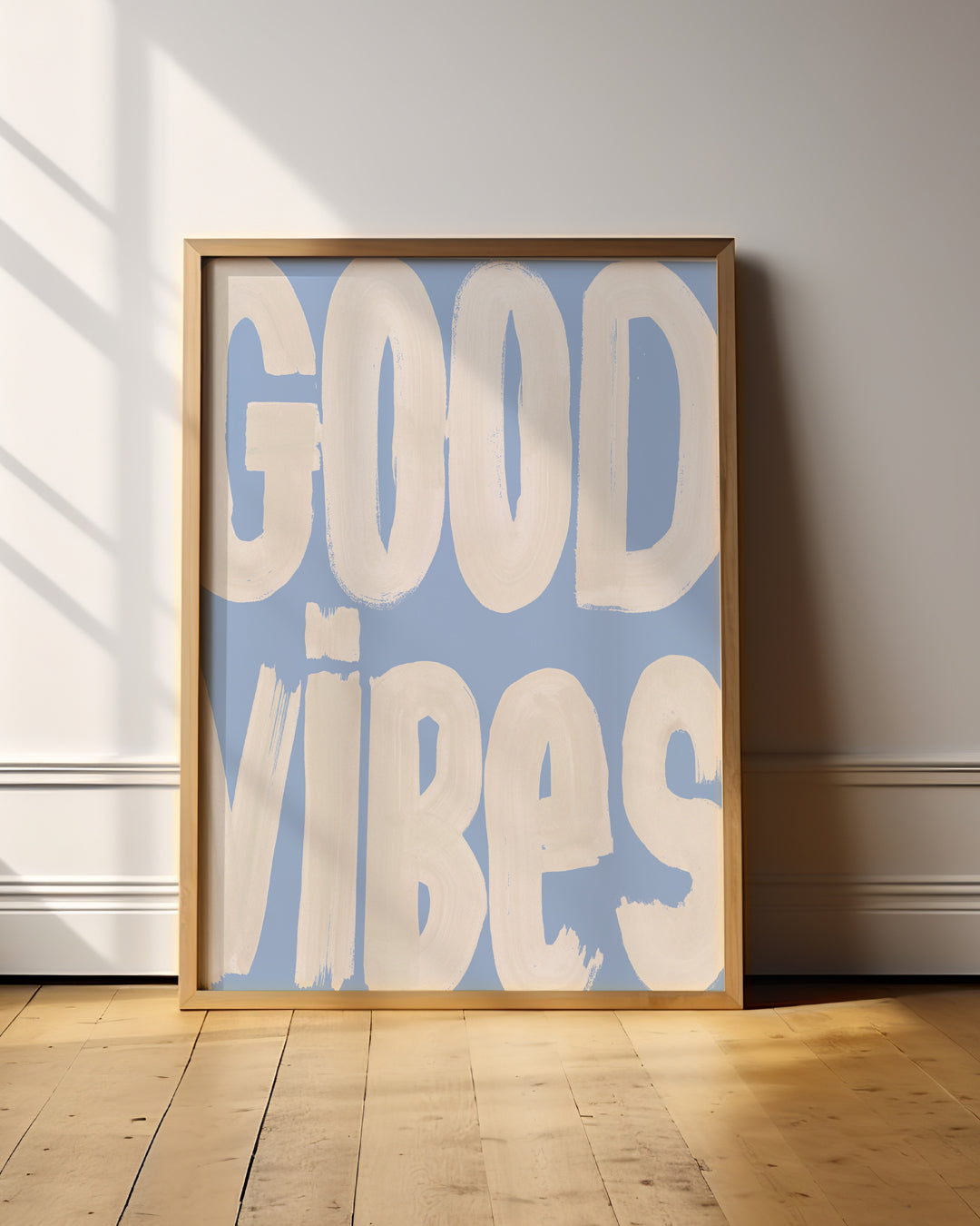 Good Vibes cream and blue typography print in a wooden frame on a sunlit wooden floor.