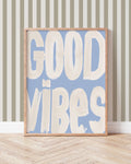 Good Vibes art print with oversized cream brush lettering on blue, displayed in a wooden frame against a striped wall.