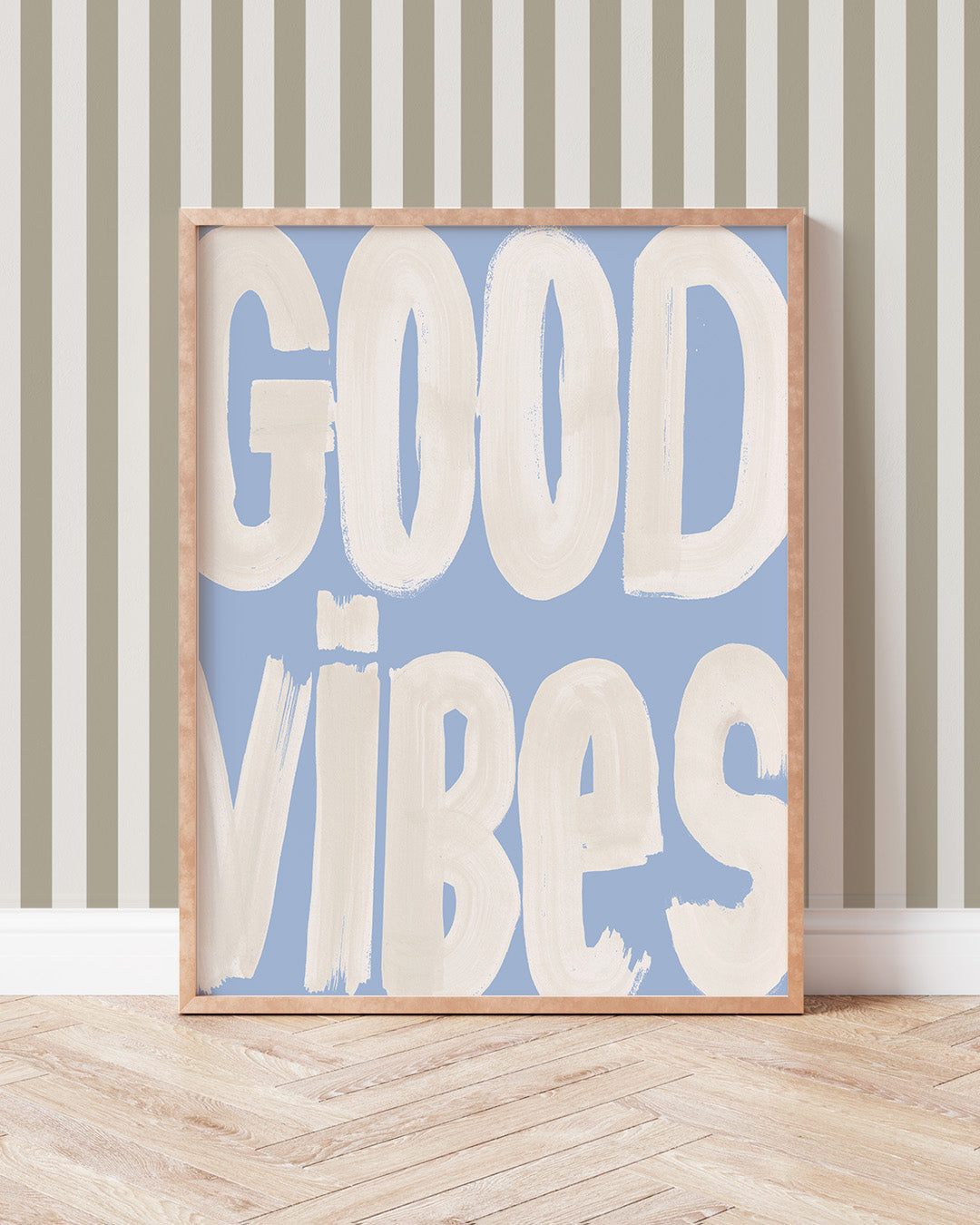 Good Vibes art print with oversized cream brush lettering on blue, displayed in a wooden frame against a striped wall.